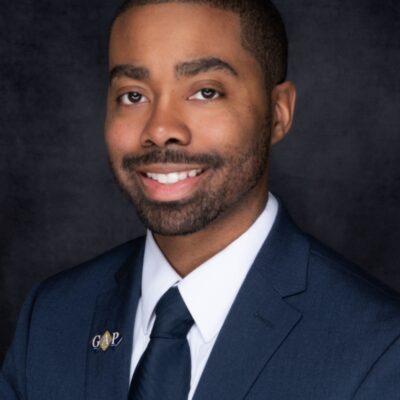 Professional portrait of a man in a navy suit and tie against a dark background.